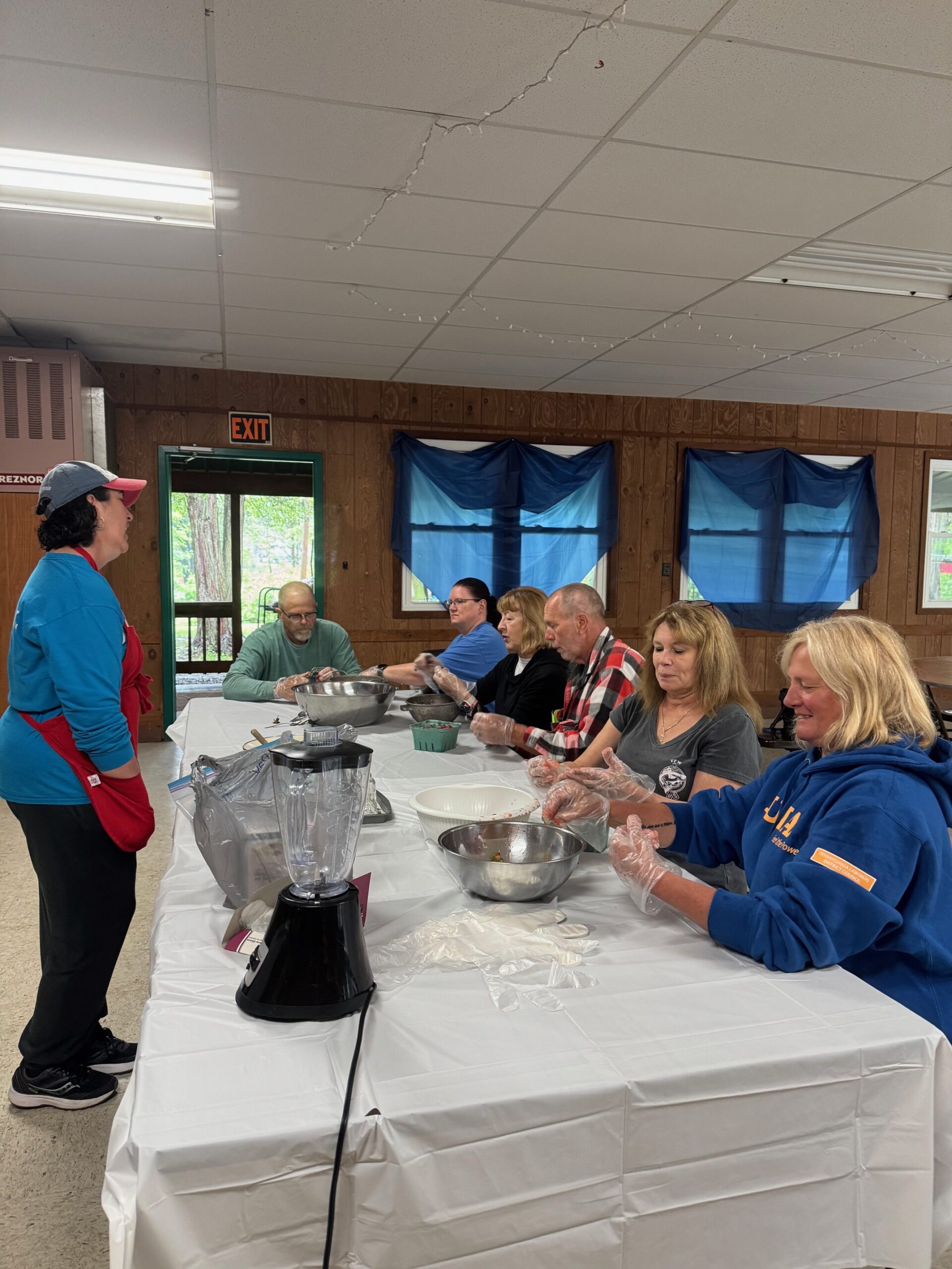 A group of seven adults sit at a long table covered with a white cloth, preparing food together. One woman stands at the end of the table, facing the group. Mixing bowls and a blender are on the table.