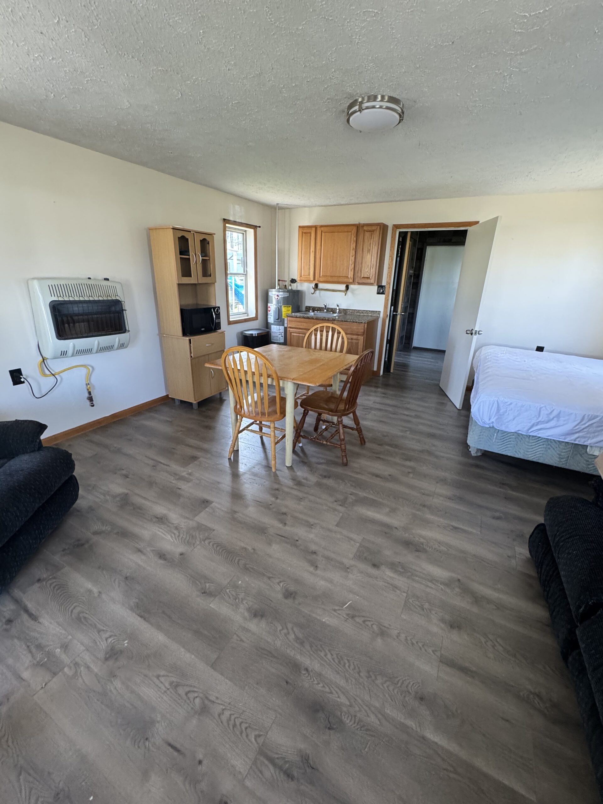 A small, sparsely furnished room with wood floors, a table and four mismatched chairs, a bed, a black armchair, a kitchenette with cabinets, and a window. The door is open, revealing another room.