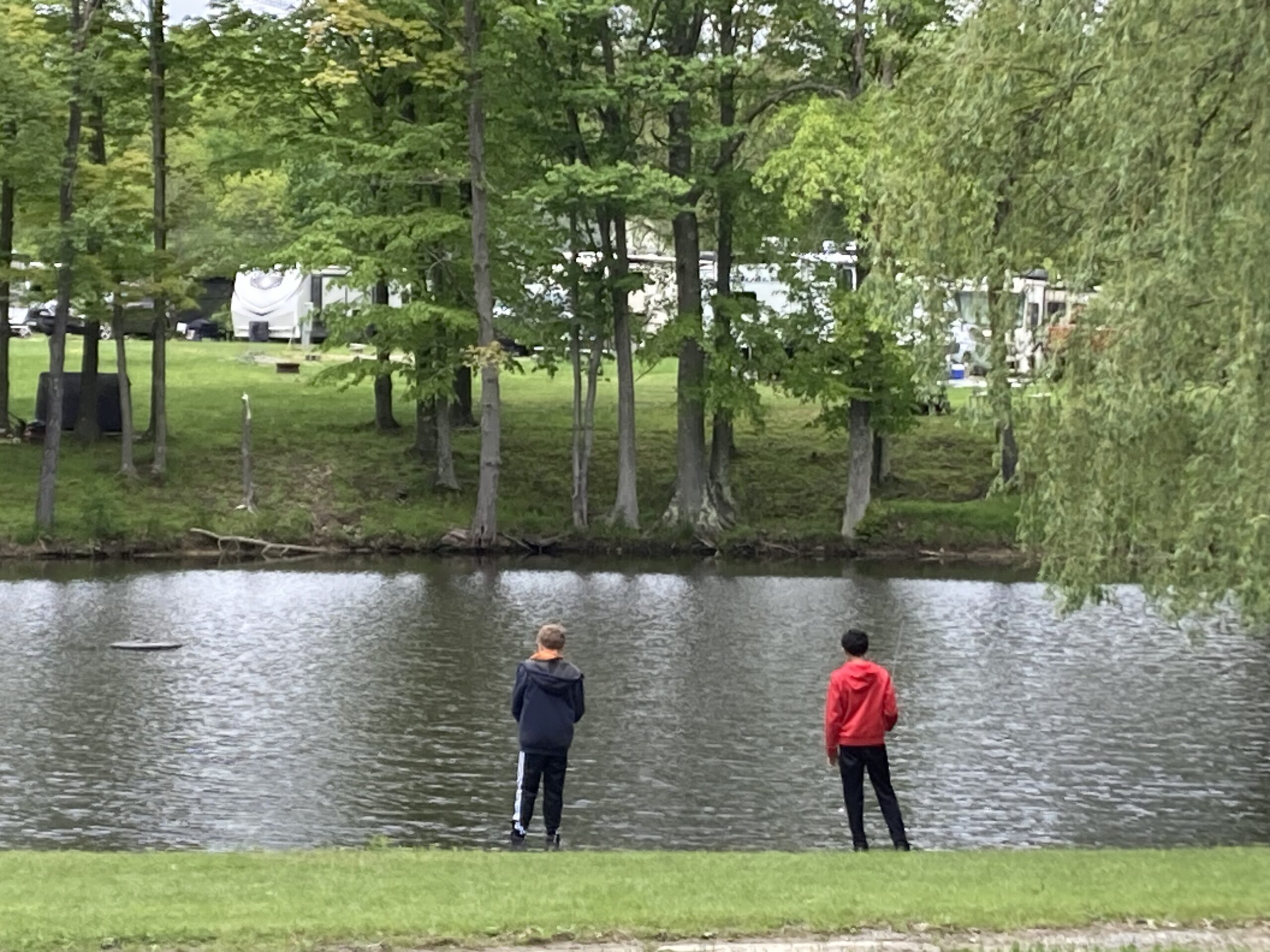 Two boys stand on the grassy bank of a lake, facing the water. Trees with green leaves line the opposite shore, and several white RVs are visible in the background, suggesting a campground setting.