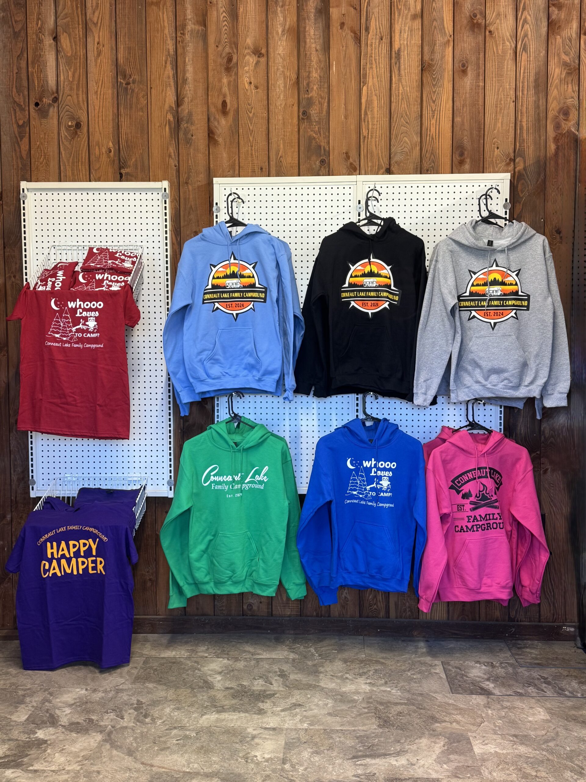 Assorted colorful hoodies and T-shirts with campground logos displayed on hangers and racks against a wooden wall and pegboard in a shop setting.