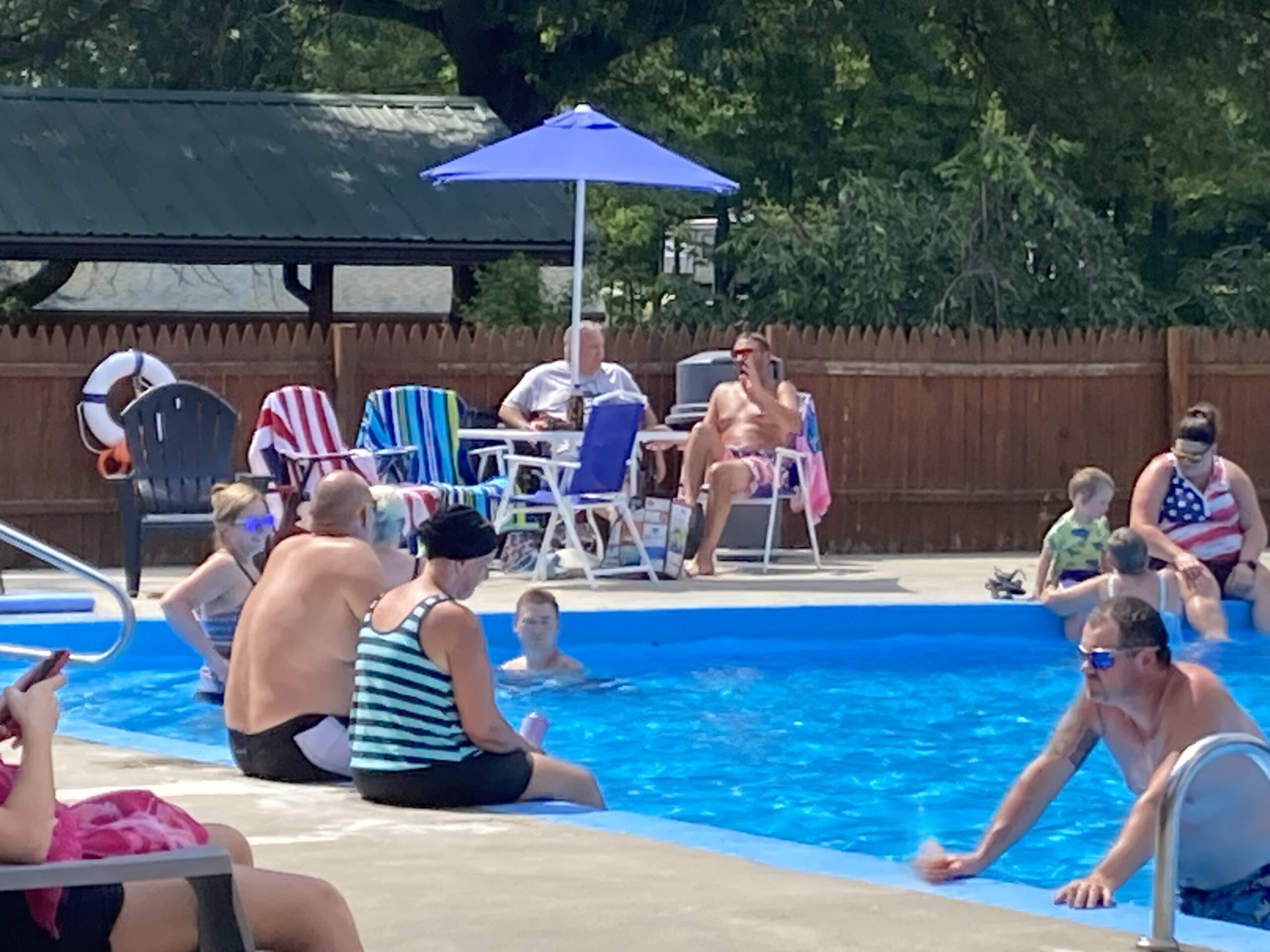 People relax by and in a swimming pool on a sunny day. Some adults sit in colorful chairs under an umbrella, while others and children swim or wade in the pool. Towels and pool toys are visible in the scene.