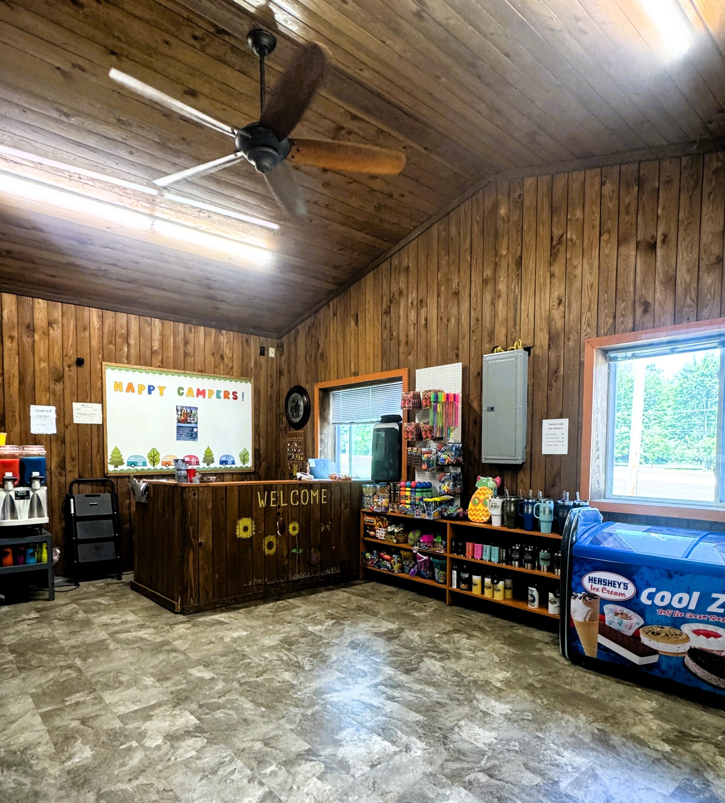 A wooden-walled camp store with a ceiling fan, a counter decorated with sunflowers and a "Happy Campers" sign, shelves stocked with snacks and supplies, and an ice cream freezer by a window.
